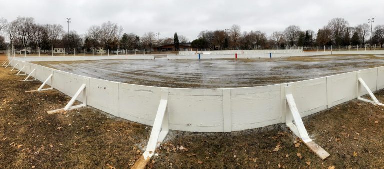 panoramic view of an ice rink with white boards in a brown field with some ice in the rink