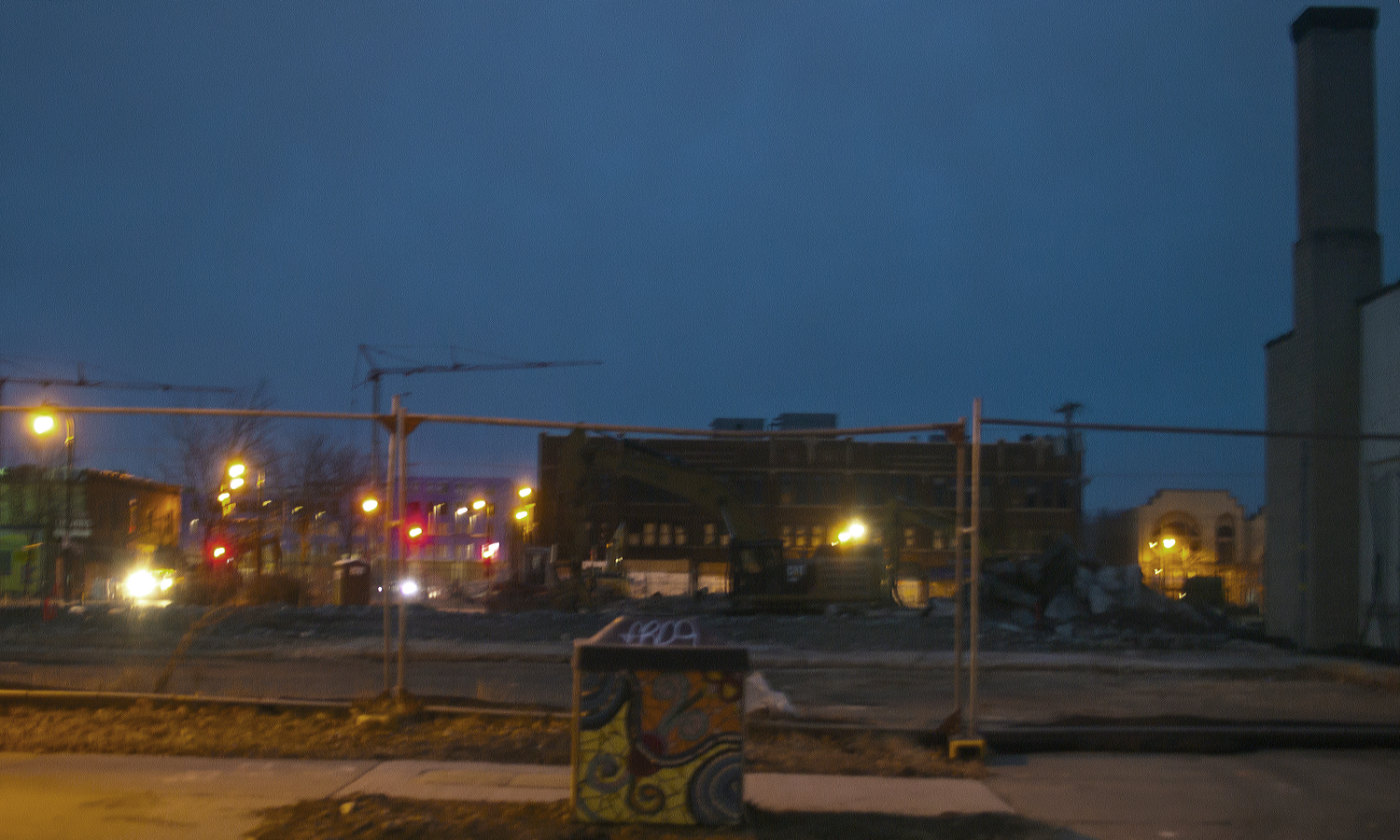 nighttime view of a fenced off lot with a row of buildings, street lights, and construction cranes in the background