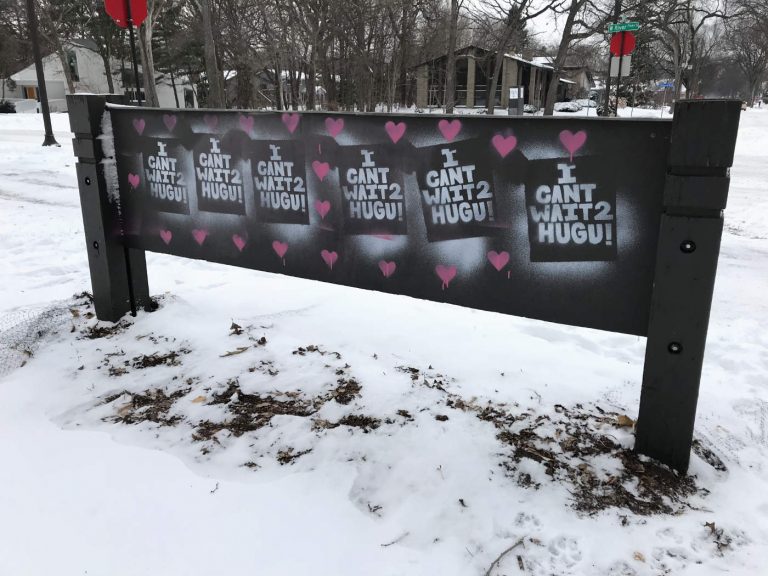 back of wide sign seen from behind in the snow with painted slogans of I CANT WAIT 2 HUG U with pink hearts