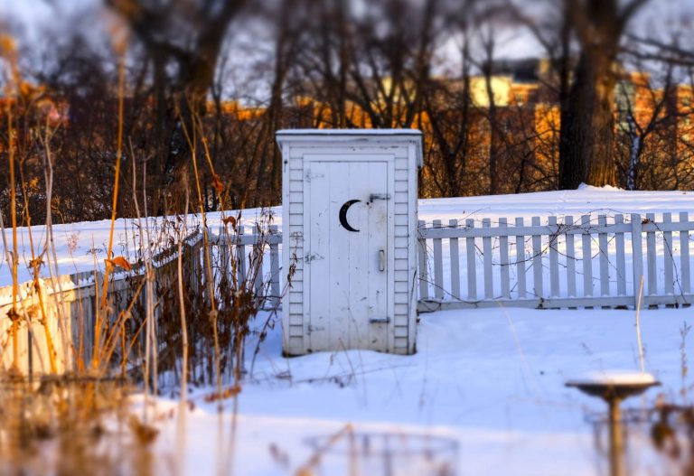 White wooden outhouse with black half-moon on door next to a white picket fence in the snow with orange sun shining on dried stalks in foreground and on blurry line of buildings in background behind trees