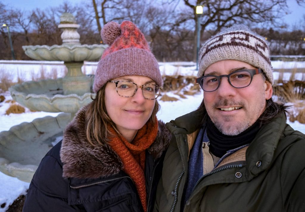woman and man with winterwear and stocking caps outside in winter with a fountain sculpture in background