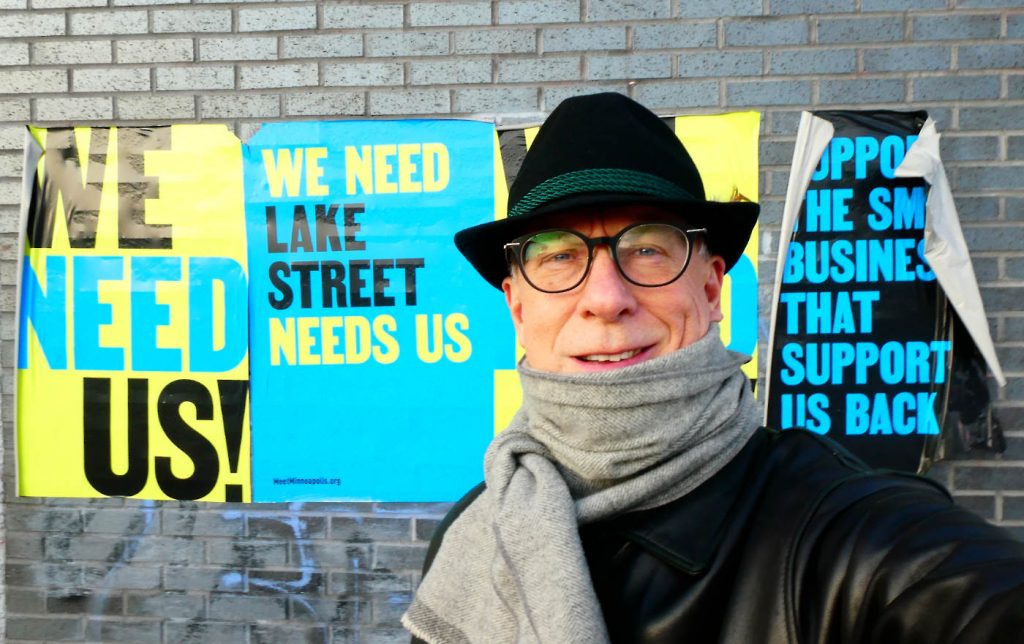 Man in winter garb looking at camera with a postered gray brick wall behind him with large text: We Need Us! We Need--Lake Street--Need Us