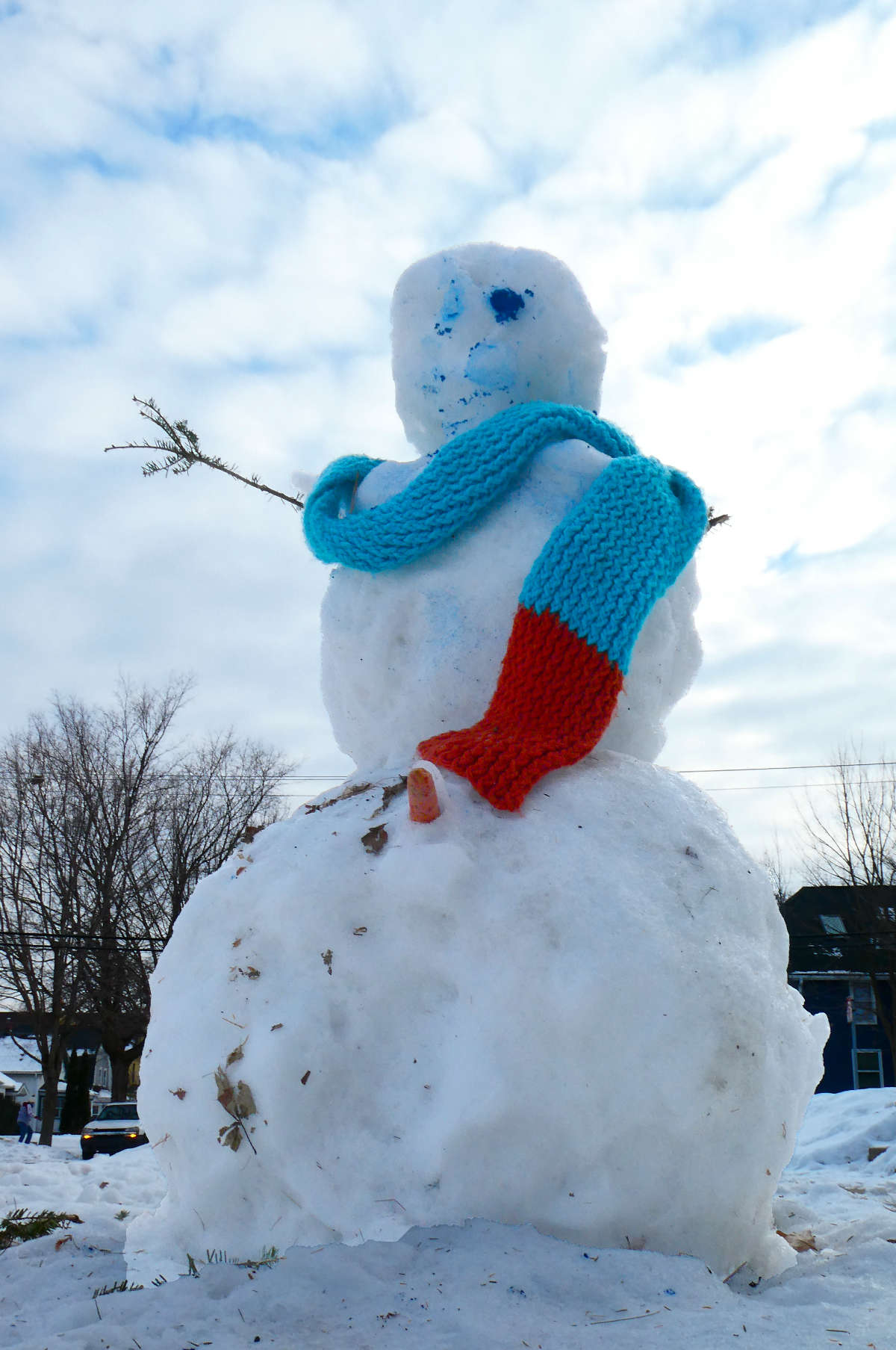 looking up at a snowman with one coal eye, one stick arm, and a blue and red scarf with a cloudy blue sky overhead