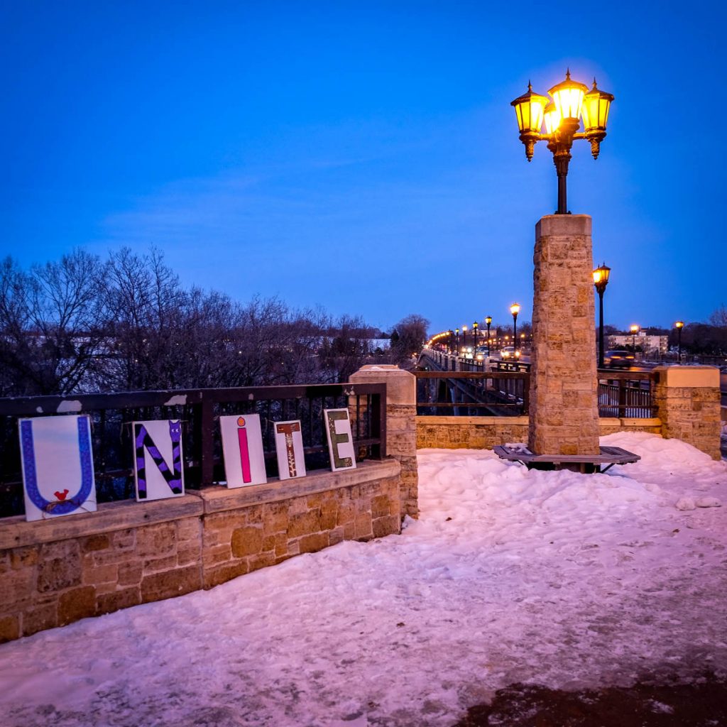 stone wall with five small signs each with a letter spelling U-N-I-T-E, with a stone column and streetlamps with receding bridge in background with deep blue sky