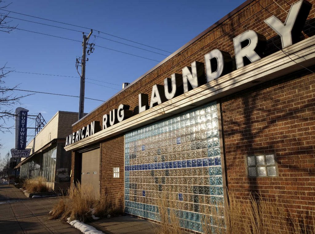single story brick storefront with colorful glass block panel and overhead metallic sign reading American Rug Laundry against a blue sky