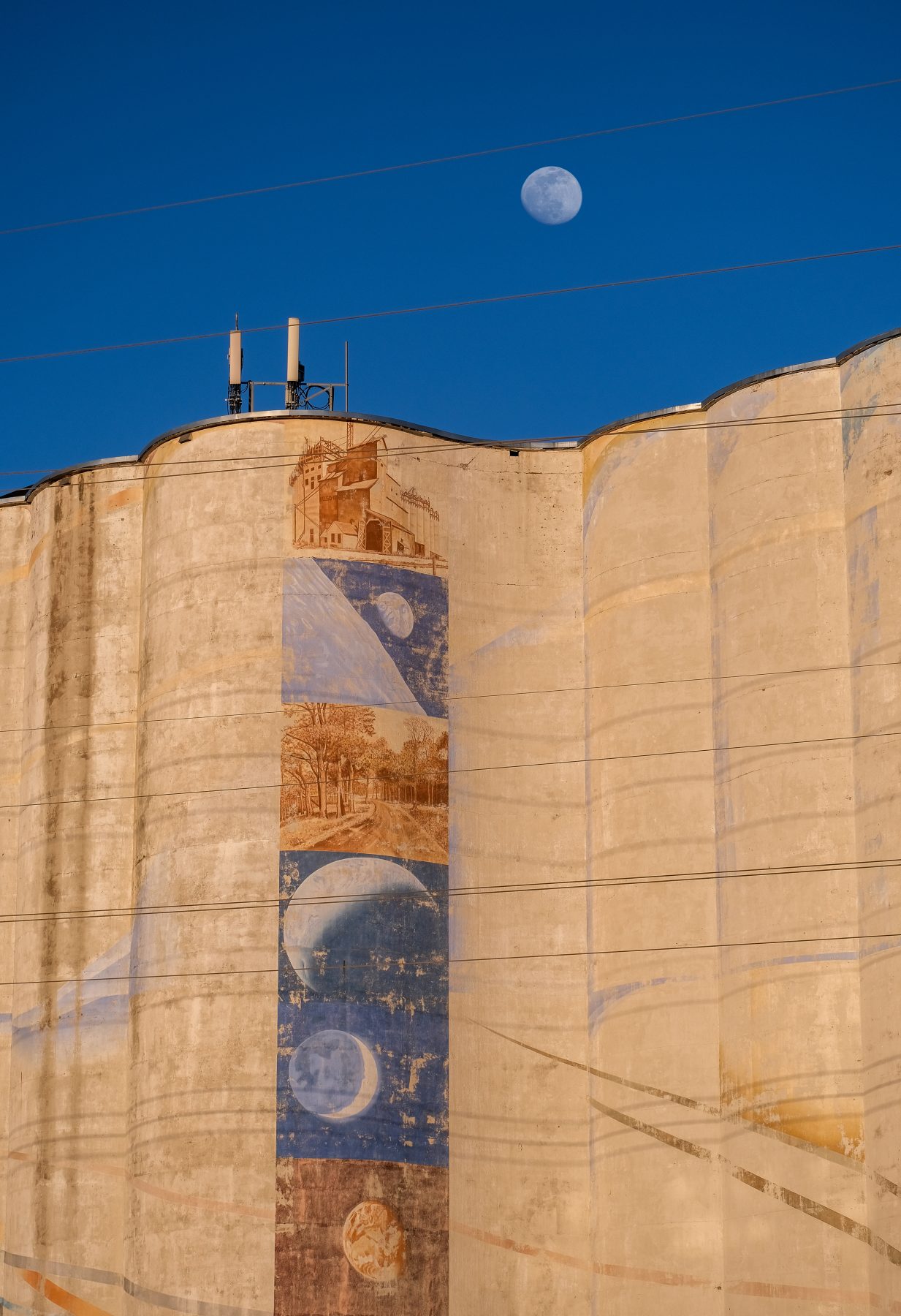 tall columns of grain elevator with vertical mural, moon appears in blue sky above