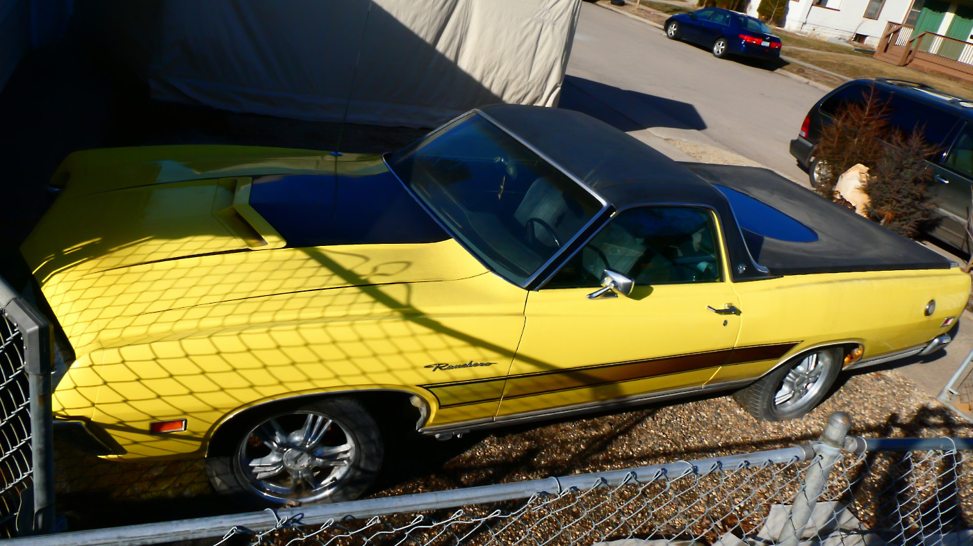 Brigh yellow classic 70s car parked with black roof and bed cover behind a fence with street in background