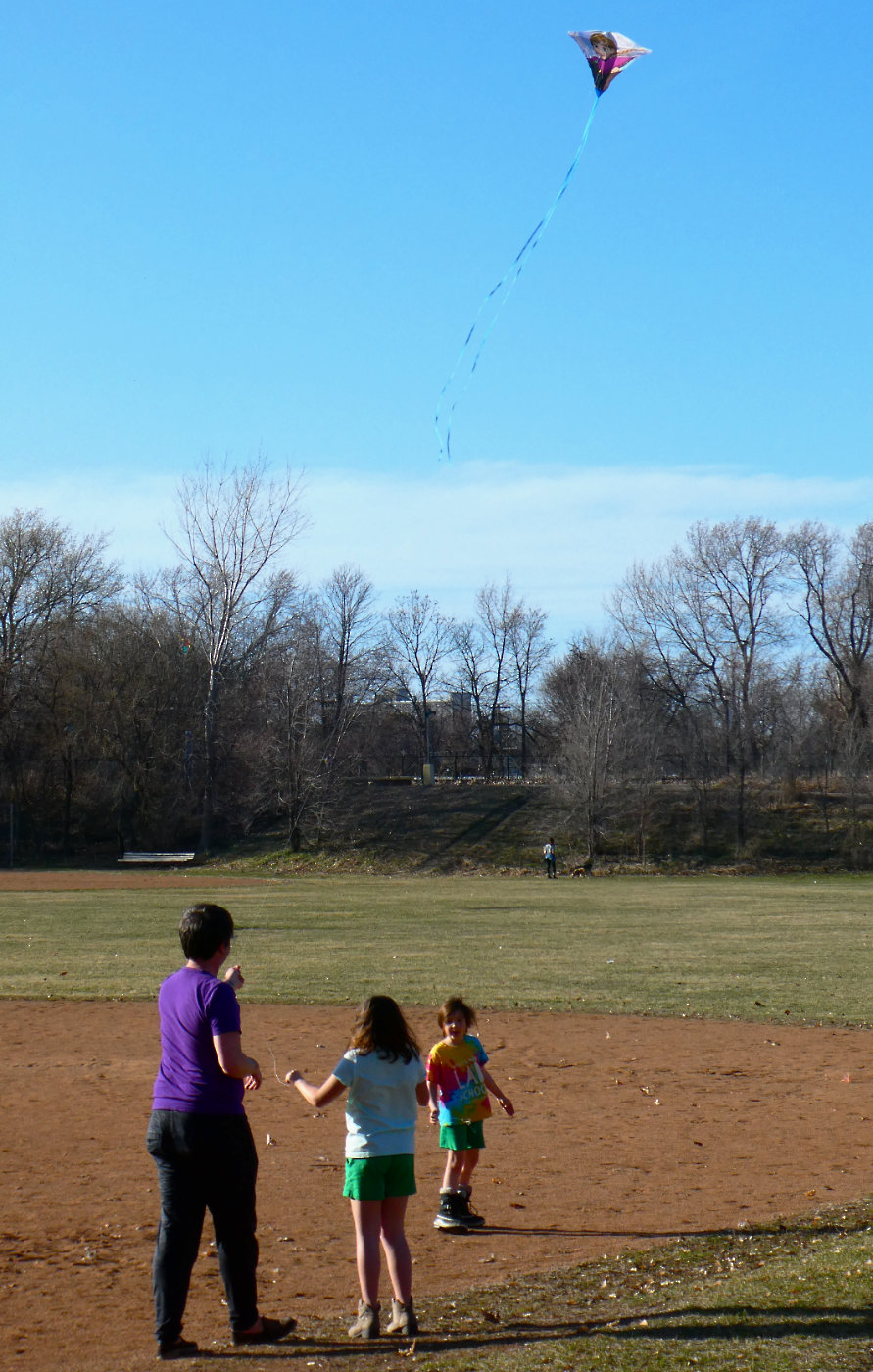 woman and two young children flying a kite on a bare baseball diamond with field and trees in background on a sunny day with deep blue sky