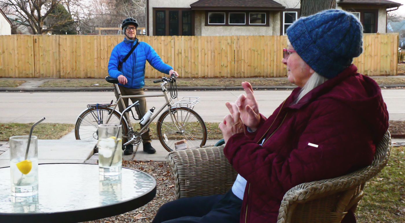Woman in cold weather garb with fingers crossed at table with two tall glasses, and a man in blue down jacket and bike helmet standing on sidewalk with a bicycle in the background