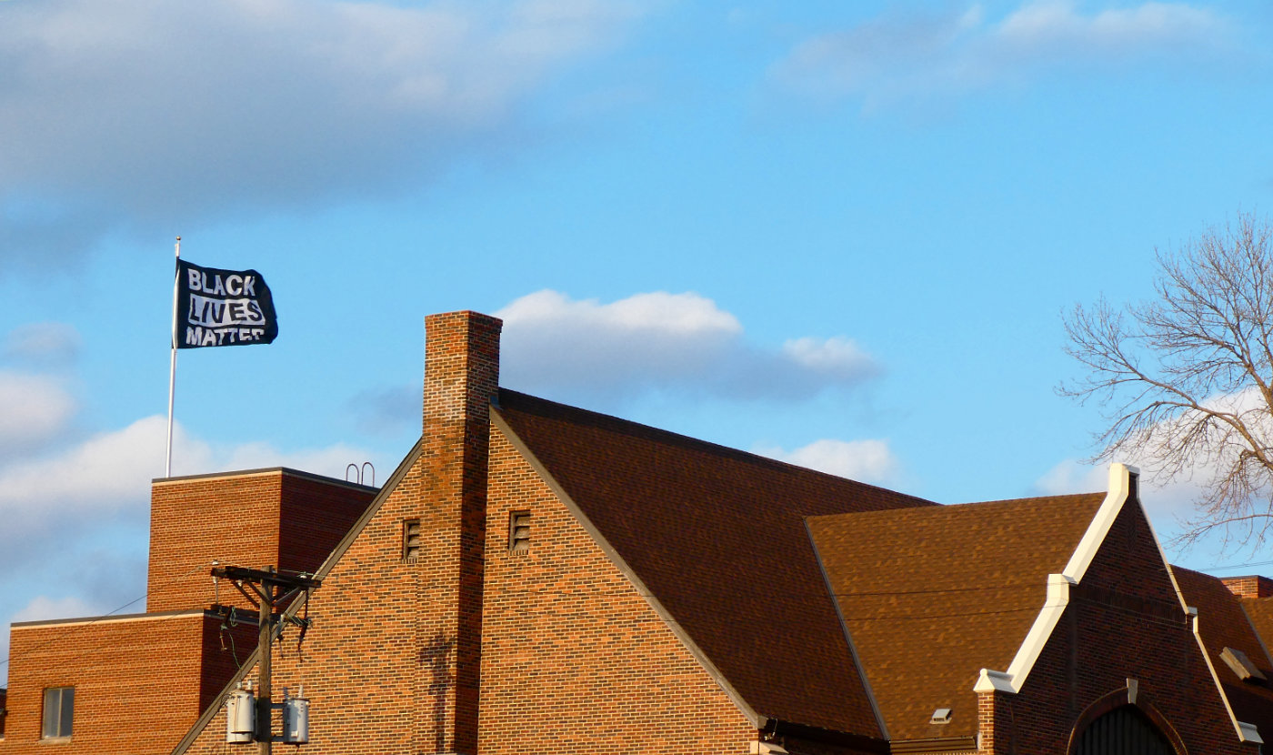 black and white Black Lives Matter flag flutters over the roof of a large brick building with chimney against a blue sky with some clouds