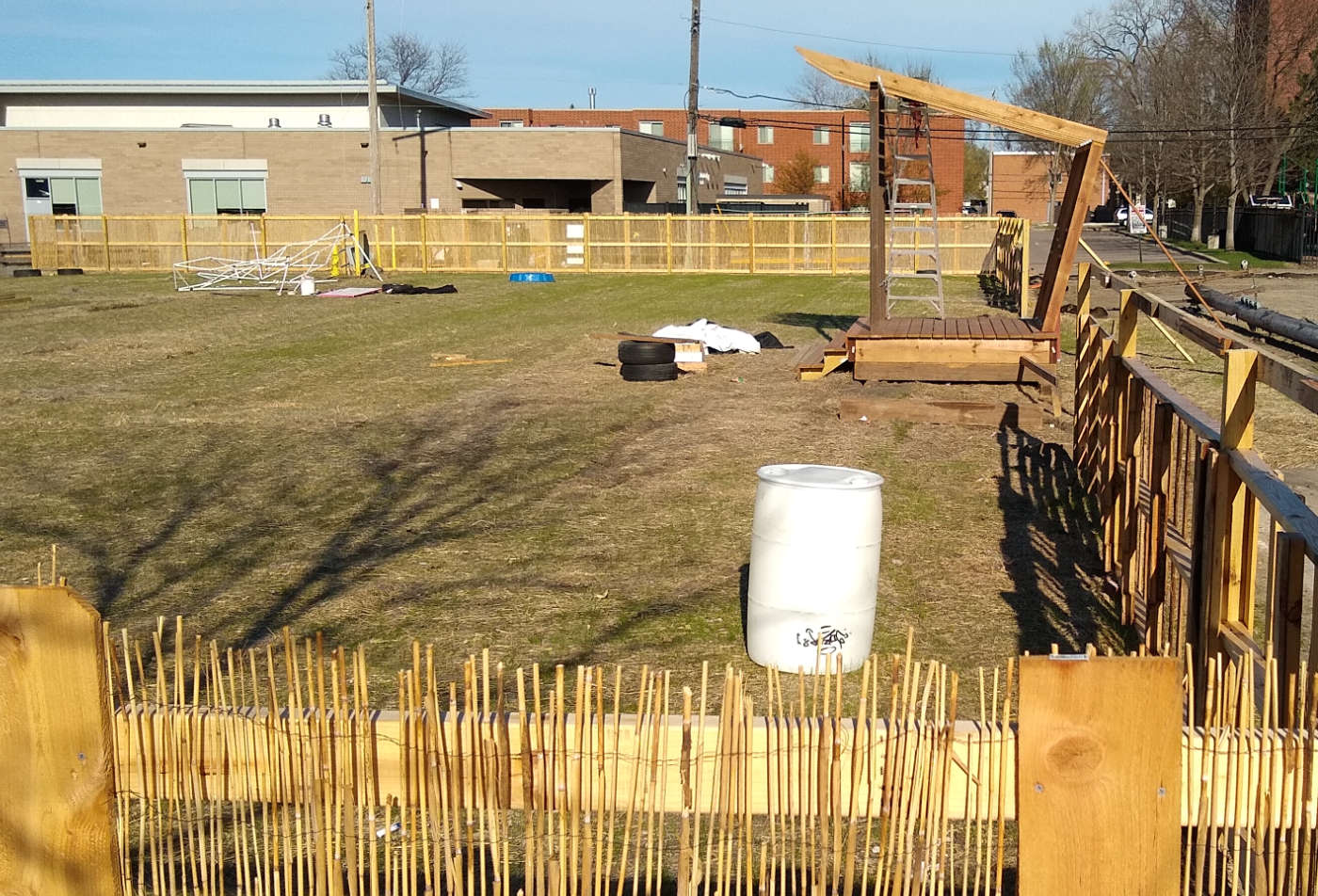 Looking over a wooden fence with cane sticks into an empty lot with a small wooden stage structure and a white barrel in foreground and other odd items on ground in distance