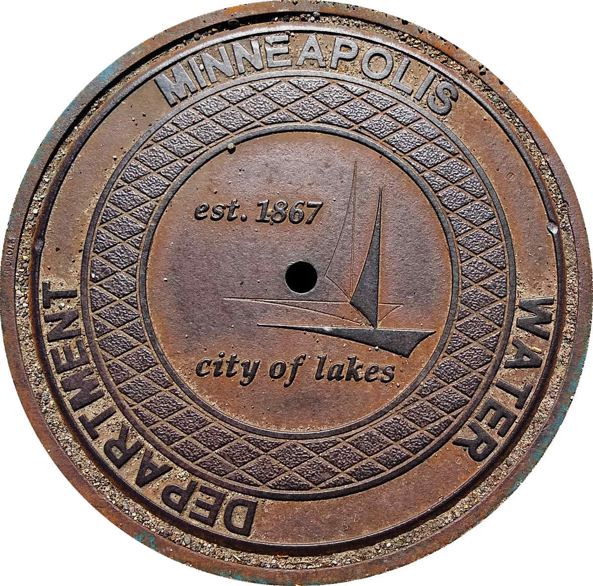 Round brown metal disk encircled with the words MINNEAPOLIS WATER DEPARTMENT with embossed designs including a sailboat silhouette in the center with the phrase "city of lakes"