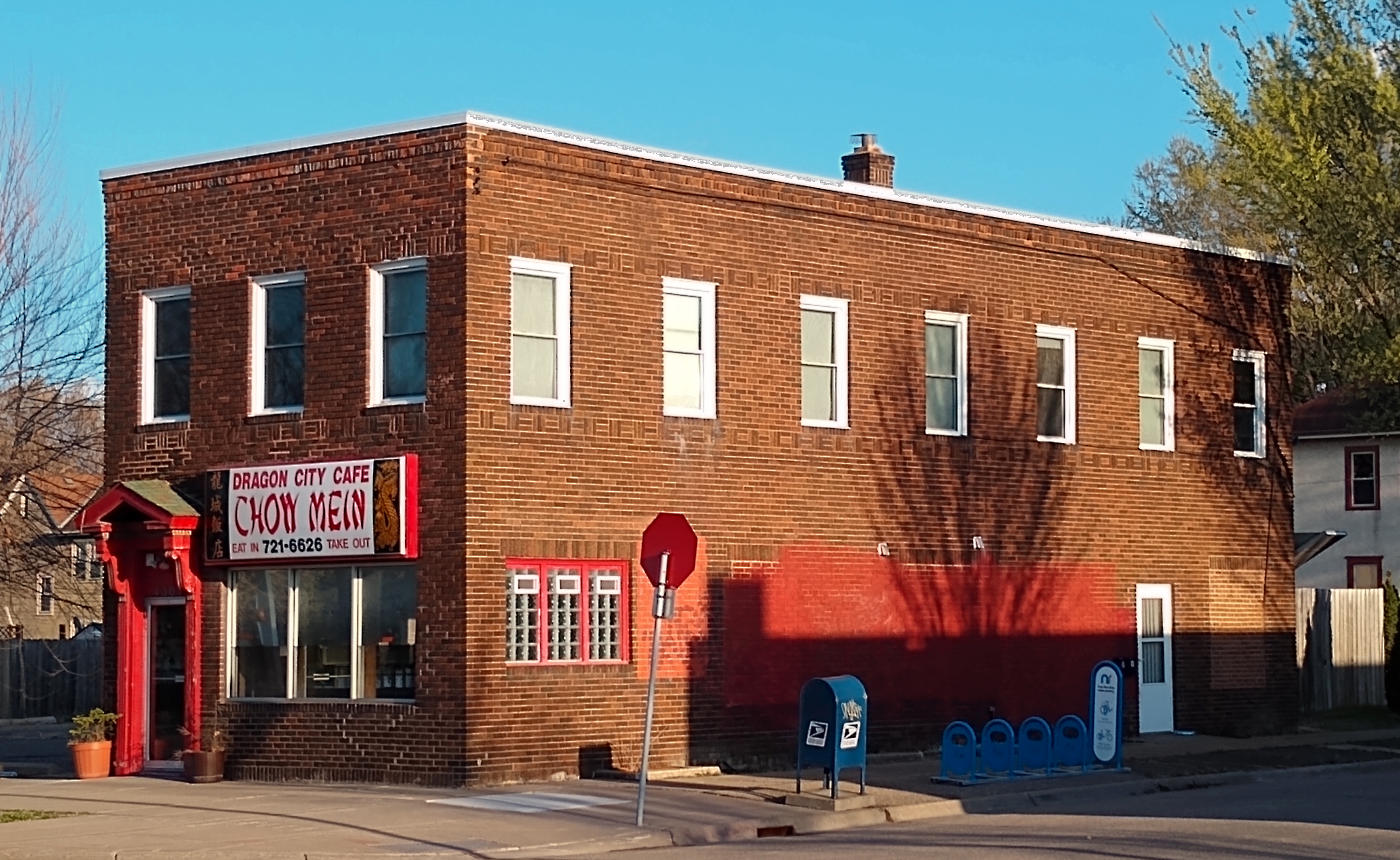 Two-story reddish-brown brick building with red trim, red and white sign over storefront reading Dragon City Cafe Chow Mein