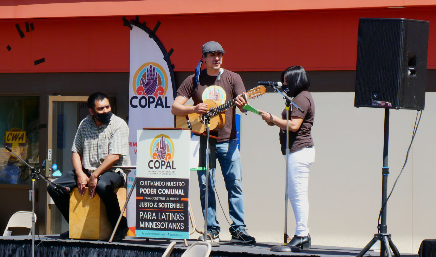 three people performing on a stage next to a sound speaker; man sitting with percussive box, man standing and playing guitar, woman singing into a microphone, with a banner and poster title COPAL.