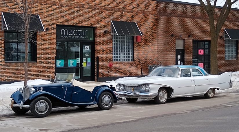 two classic cars parked on street, one a convertible with a brick storefront in background