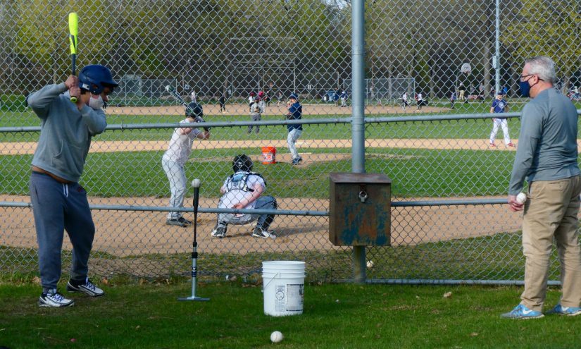 Looking over a baseball diamond from behind the fenced backstop with players on field, on deck batter with T-ball in foreground, and adult coach on the side